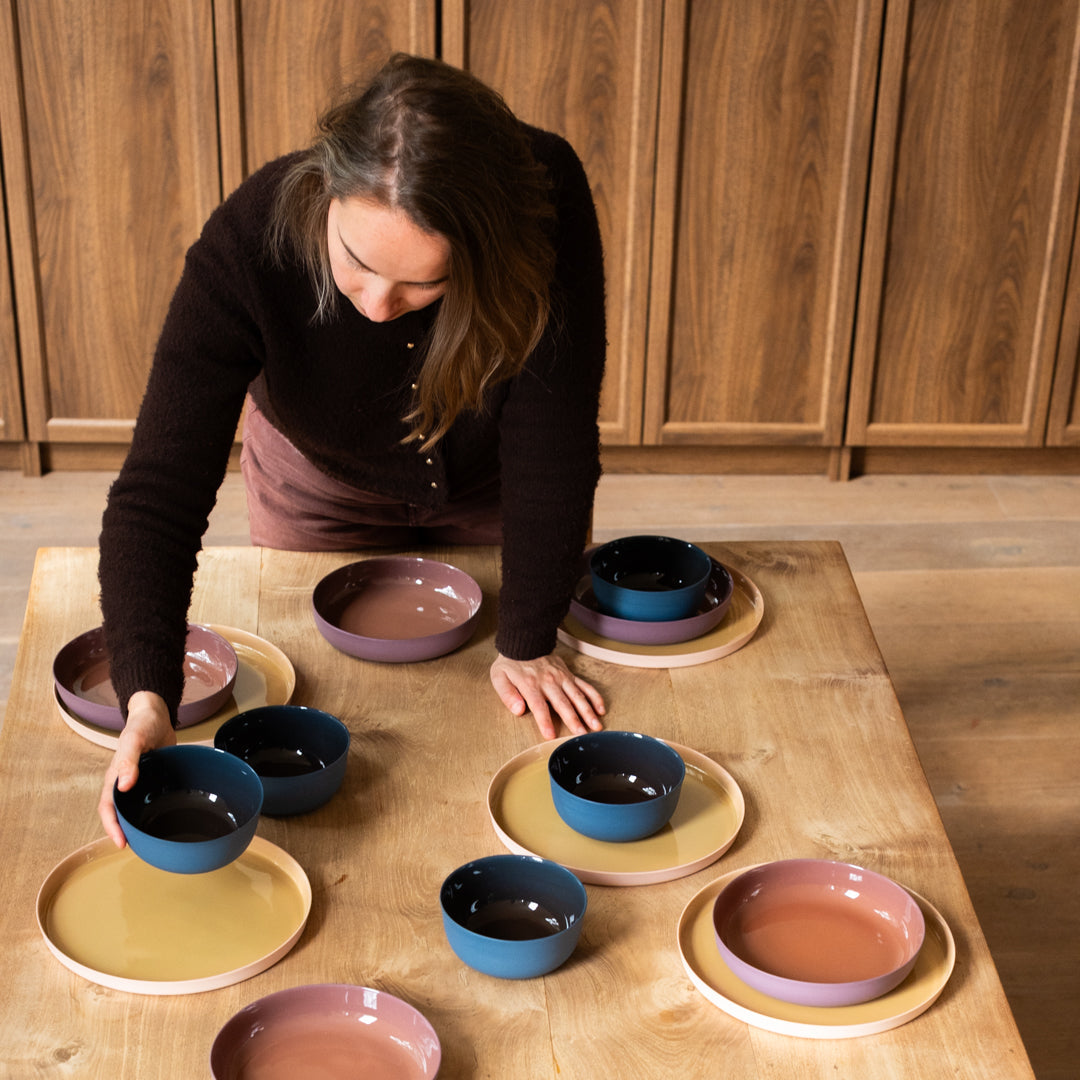 A woman arranges colorful ceramic bowls and plates on a light wood table in a kitchen setting with wooden cabinets in the background.