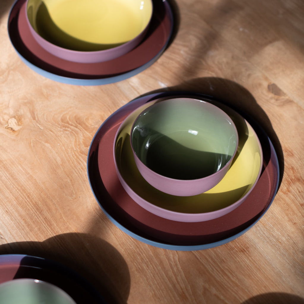 Colorful stacked ceramic bowls and plates in yellow, green, pink, and dark blue, arranged on a warm wooden tabletop with natural sunlight and shadows.