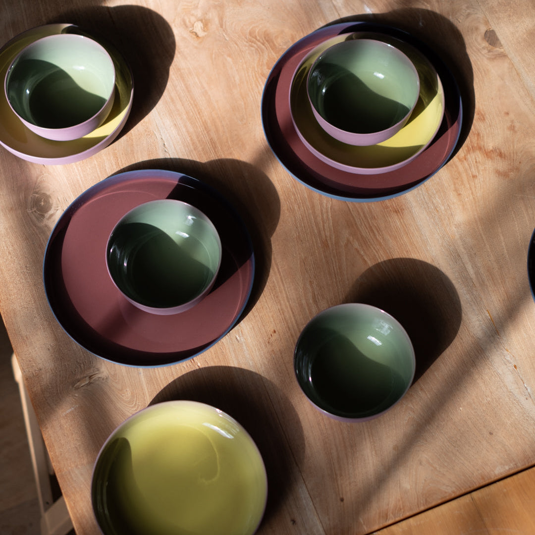 A collection of hand-painted ceramic tableware, including plates, bowls, and cups, arranged on a wooden table under dramatic natural sunlight and shadows.