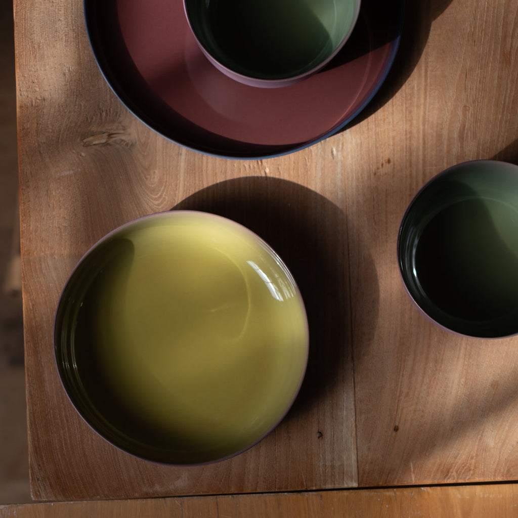 A top-down view of a wooden dining table set with ceramic bowls and plates in muted earthy tones, including a large pale yellow-green bowl, a dark green cup, and a reddish-brown plate.