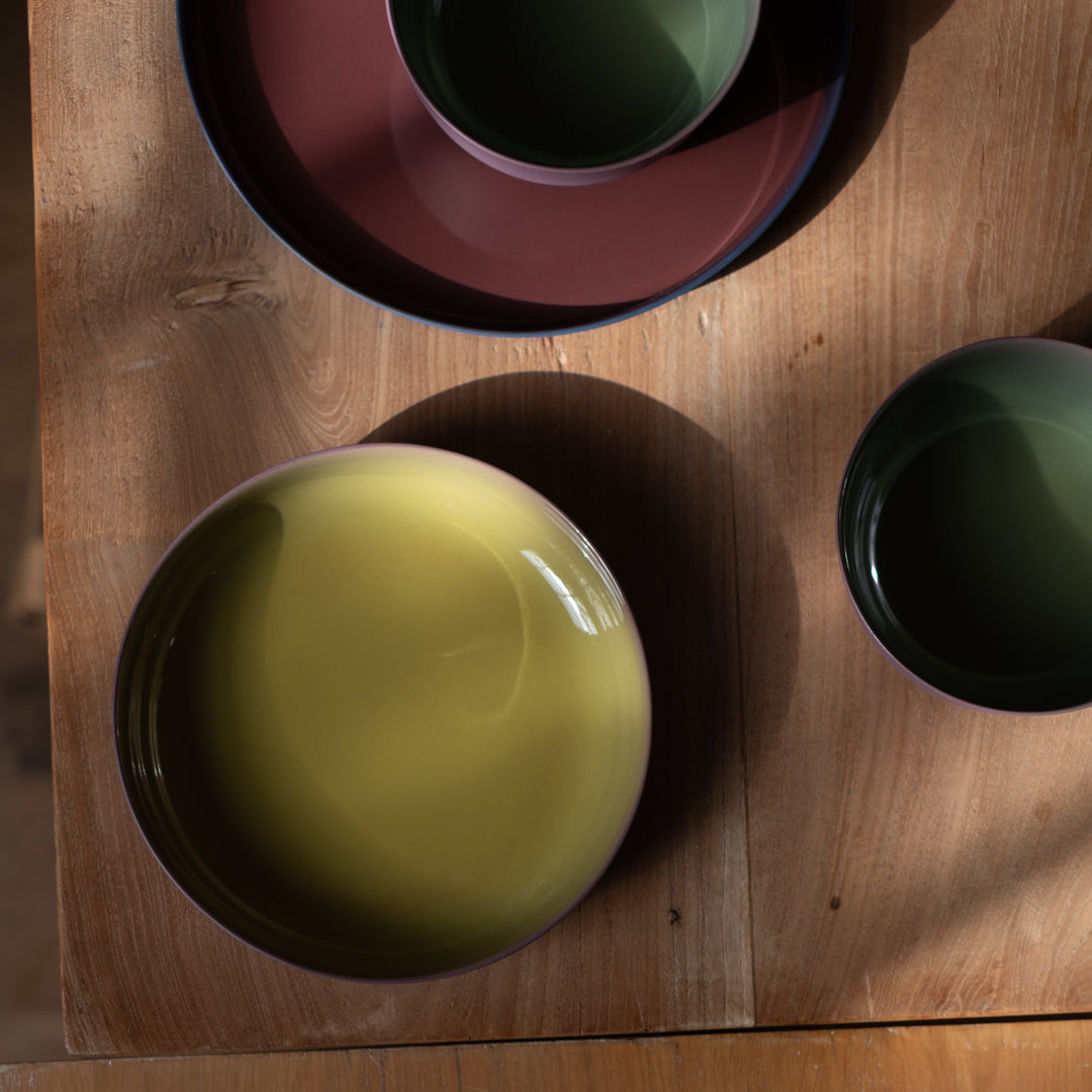A top-down view of a wooden dining table set with ceramic bowls and plates in muted earthy tones, including a large pale yellow-green bowl, a dark green cup, and a reddish-brown plate.