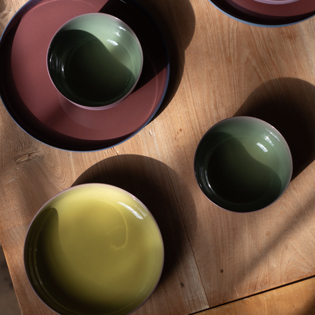 A set of ceramic bowls and plates in muted earthy tones, including olive green, mustard yellow, dusty rose, and dark purple, arranged on a wooden table with dramatic natural shadows.